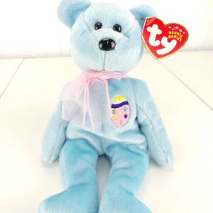 May include: A light blue Beanie Baby bear plush toy with a pink ribbon around its neck. The bear has a colorful Easter egg design on its chest and a red heart-shaped tag.