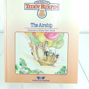 May include: A book cover for "The Airship" from the "The World of Teddy Ruxpin" series. The cover features a cartoon illustration of a hot air balloon shaped like a ship with Teddy Ruxpin and his friends on board. The book is published by Worlds of Wonder.