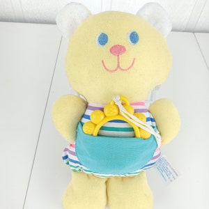 May include: A yellow plush teddy bear wearing a striped dress with a blue pocket. The bear is holding a yellow plastic teether with a rope attached.