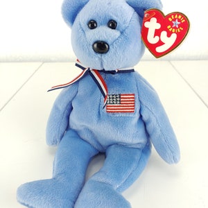 May include: A light blue plush bear with a black nose and eyes. It has a small American flag patch and a red heart-shaped tag. A red, white, and blue ribbon is tied around its neck. The bear is a collectible item.