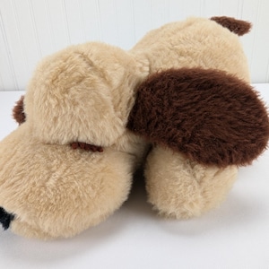 Puede incluir: Un juguete de peluche para perro de color beige y marrón con orejas caídas. El juguete está hecho de un material suave y esponjoso y tiene una nariz marrón.