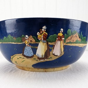 May include: A large blue ceramic bowl with a hand-painted scene of women in traditional Dutch clothing walking along a path by a body of water. The bowl has a light brown rim.