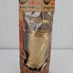 Puede incluir: Un molde para hacer donas Mirro vintage en su embalaje original. El molde para hacer donas de metal dorado está en una bolsa de plástico transparente. La caja es marrón con letras rojas e imágenes de donas.