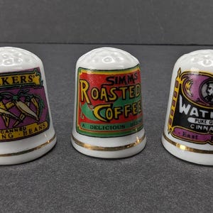 Può includere: Tre ditiali in ceramica bianca con bordi dorati. Uno recita "SHAKERS CANNED STRING BEANS", un altro "SIMMS ROASTED COFFEE" e l'ultimo "WATKINS PURE GROUND CINNAMON East Indies". I ditiali hanno etichette colorate.