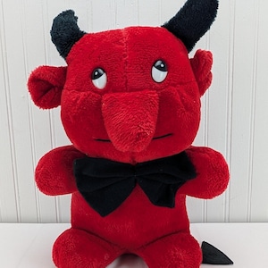 Puede incluir: Un muñeco de diablo rojo de peluche con cuernos, orejas y pajarita negros. El muñeco tiene una nariz larga, brazos pequeños y una cola pequeña. Tiene ojos blancos y está sobre una superficie blanca.