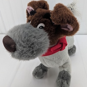 May include: A soft toy dog with a brown head and ears, a grey snout, and a red scarf. The dog's body is grey, and it has black eyes. The toy is designed to resemble a cartoon dog.