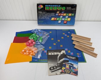 Może przedstawiać: Zestaw do gry w Scrabble Rebus w stylu vintage. Pudełko jest granatowe z kolorową grafiką i tekstem. Gra zawiera niebieską planszę, drewniane uchwyty na kafelki i różne kolorowe elementy gry w plastikowych woreczkach. Gra jest dla dorosłych i dzieci w wieku od 8 lat.