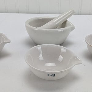 以下が含まれることがあります： 白のセラミック製実験器具のコレクション。乳鉢と乳棒、注ぎ口付きの小さなボウルが3つ含まれています。乳鉢と乳棒が中央にあり、ボウルがその周りに配置されています。各ボウルには小さな濃い青色のシンボルが付いています。