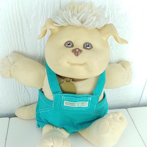 Puede incluir: Un juguete de peluche de perro Cabbage Patch Kids con un mono azul, un collar marrón y una etiqueta dorada. El perro tiene una melena blanca y esponjosa, ojos marrones y una nariz marrón.