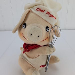 May include: A plush pig toy dressed as a chef, wearing a white chef's hat with "Chef Pigerre" embroidered in red. The pig has a red scarf and is holding a metal whisk. The toy has a smiling face and is wearing a white apron.