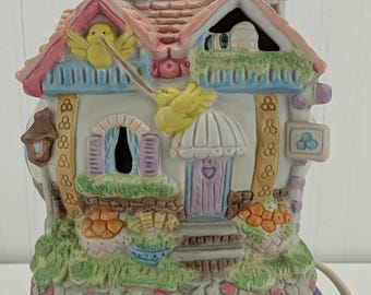 May include: A ceramic house-shaped figurine with a pink roof, blue chimney, and a sign that says "BAKERY". The house has a white facade with a purple door, and is decorated with yellow chicks and floral accents. The base is green and textured.
