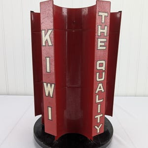 Puede incluir: Un expositor vintage rojo y negro de Kiwi The Quality. La pantalla tiene una forma curva con las palabras "KIWI" y "THE QUALITY" en letras blancas. Se asienta sobre una base circular negra.