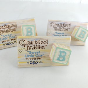 Könnte beinhalten: Drei Schubladengriffe mit einem pastellblauen, gelben und rosafarbenen Block mit dem Buchstaben "B" in hellblau. Die Verpackung lautet "Cherished Teddies" "Sweet Little One" Drawer Pull by ENESCO.