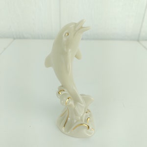 May include: A white porcelain figurine of a dolphin leaping out of the water. The dolphin is painted with gold accents and has a detailed design.
