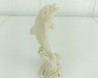以下が含まれることがあります： 水から飛び出すイルカの白い磁器の置物。イルカは金色のアクセントで描かれており、細部までこだわったデザインです。