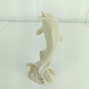 May include: A white porcelain figurine of a dolphin leaping out of the water. The dolphin is painted with gold accents and is set on a base with a wave design.