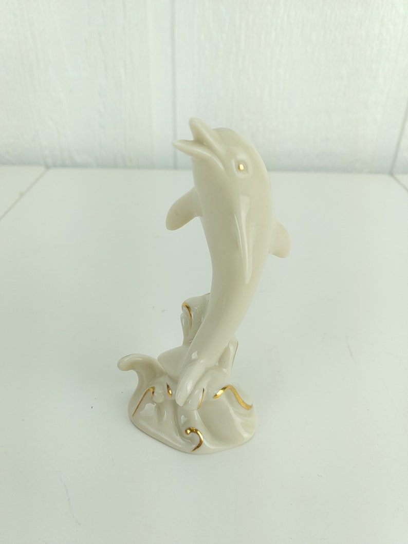 May include: A white porcelain figurine of a dolphin leaping out of the water. The dolphin is painted with gold accents and is set on a base with a wave design.