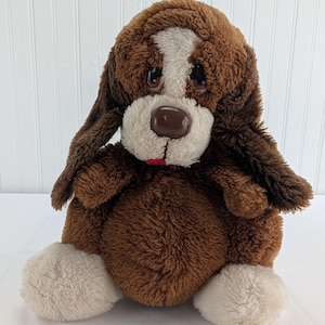 May include: A plush brown and white puppy dog stuffed animal with long floppy ears, a white patch on its forehead, and a red stitched mouth. The puppy has a brown plastic nose and sad-looking eyes.