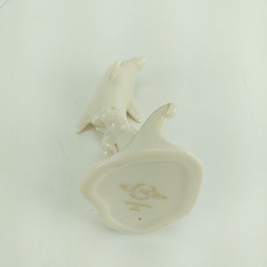 May include: A white porcelain figurine of two dolphins, handcrafted in China by Lenox. The figurine is in the shape of two dolphins swimming together, with the smaller dolphin riding on the back of the larger dolphin. The figurine is marked with the Lenox logo and the words "Handcrafted in China".