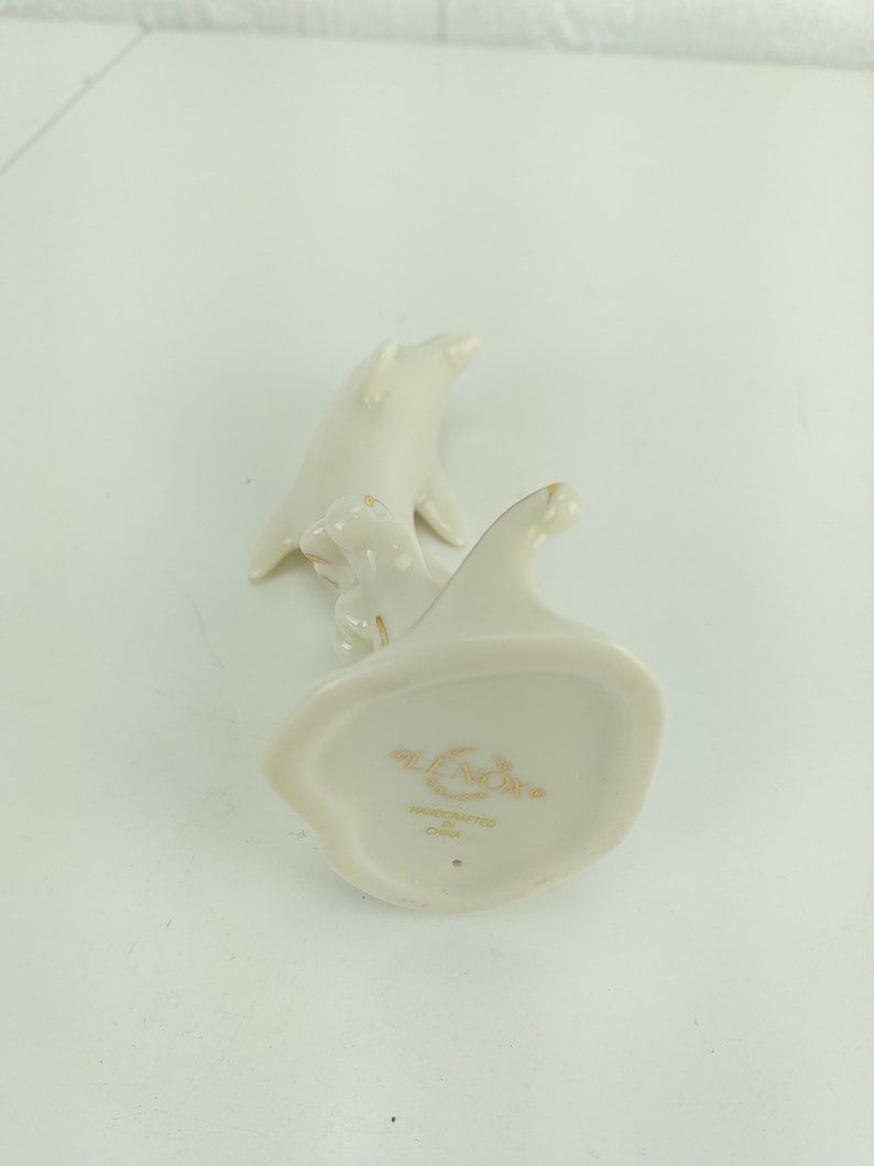 May include: A white porcelain figurine of two dolphins, handcrafted in China by Lenox. The figurine is in the shape of two dolphins swimming together, with the smaller dolphin riding on the back of the larger dolphin. The figurine is marked with the Lenox logo and the words "Handcrafted in China".