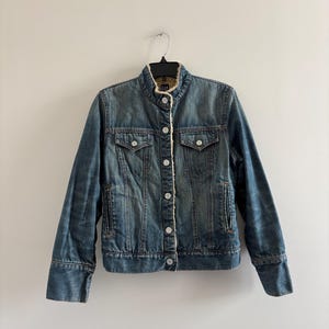 Könnte beinhalten: Eine blaue Jeansjacke mit Stehkragen und beigem Sherpa-Futter. Die Jacke verfügt über eine Knopfleiste vorne, zwei Brusttaschen mit geknöpften Klappen und zwei Seitentaschen. Das Gap-Markenetikett ist am inneren Kragen sichtbar.