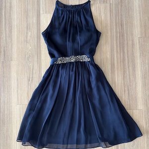 May include: A navy blue sleeveless dress featuring a high halter neckline with gathered fabric details. The waist is accented by a horizontal band embellished with clear, sparkling crystals and beads, leading into a pleated, knee-length skirt.