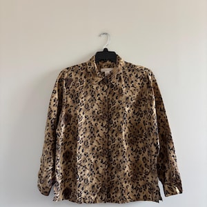 Könnte beinhalten: Langärmliges Hemd mit Leopardenmuster. Das Hemd hat einen klassischen Kragen und geknöpfte Manschetten. Das Tiermusterdesign umfasst Brauntöne und Schwarz.