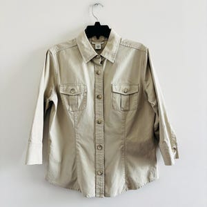 May include: A tan button-down shirt with two chest pockets and three-quarter sleeves. The shirt is hanging on a black hanger against a white background. The shirt has a collared neckline and button closures down the front.