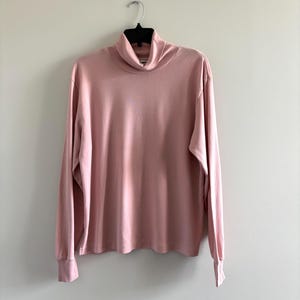 May include: A pale pink turtleneck top with long sleeves, hanging on a black hanger. The top features a high, folded collar and ribbed cuffs. The material looks soft and comfortable.
