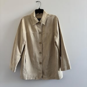 May include: A tan-colored jacket with a collared neckline and button-down closure. The jacket has long sleeves and is hanging on a black hanger against a white wall. The jacket appears to be made of a soft, suede-like material.