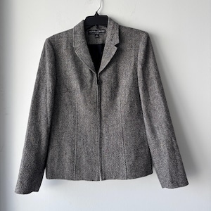 May include: A gray tweed blazer with a notched collar and a full-length zipper. The jacket is hanging on a black hanger against a white wall. The blazer has a tailored fit and a classic design.