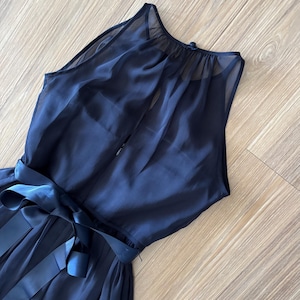 May include: A sleeveless navy blue dress featuring a sheer overlay on the bodice, a high neckline with a button closure at the back, and a matching satin ribbon tie at the waist. The skirt is gathered and flows from the waistline downward.