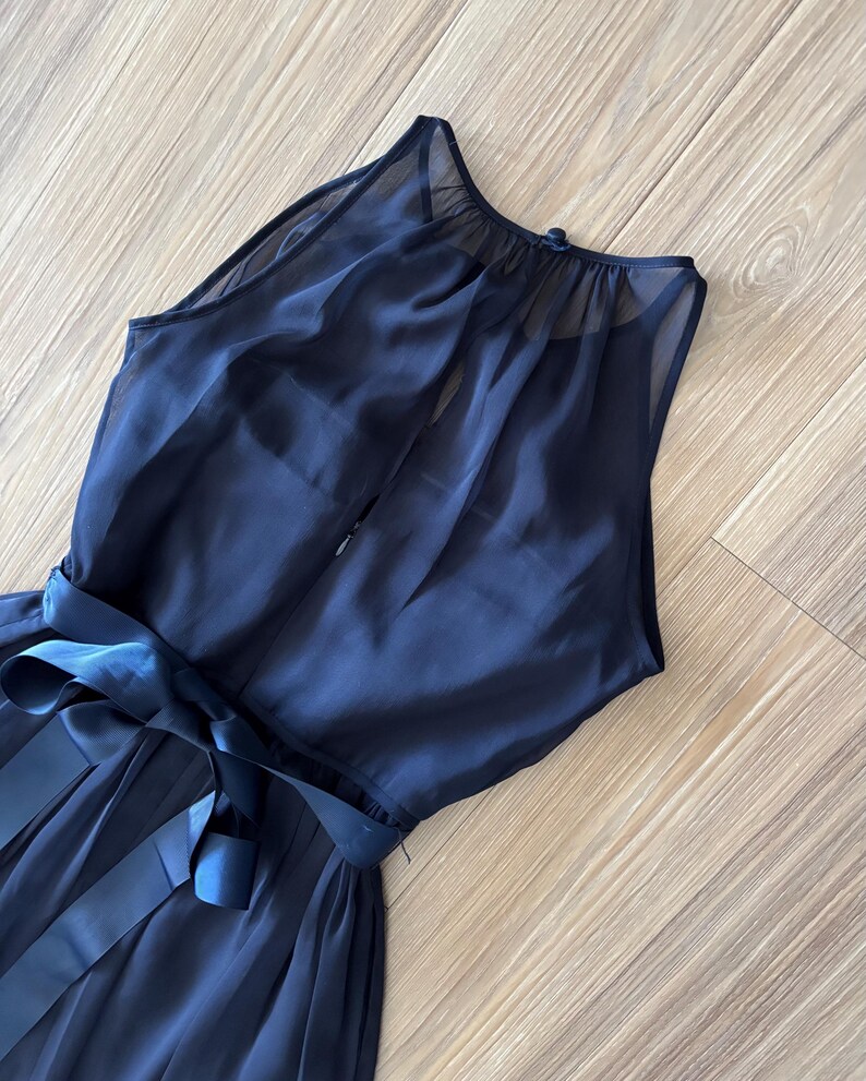 May include: A sleeveless navy blue dress featuring a sheer overlay on the bodice, a high neckline with a button closure at the back, and a matching satin ribbon tie at the waist. The skirt is gathered and flows from the waistline downward.