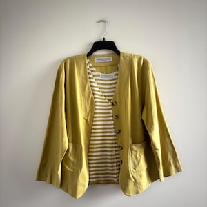 May include: A mustard yellow jacket with a V-neck and two front pockets, paired with a striped shirt. The jacket has button closures and three-quarter sleeves. The shirt has a white and olive green striped pattern.