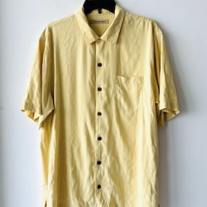 May include: A pale yellow short-sleeved shirt with a button-down front and a collared neckline. It features a single chest pocket and dark brown buttons. The shirt is suspended on a black hanger against a white backdrop.