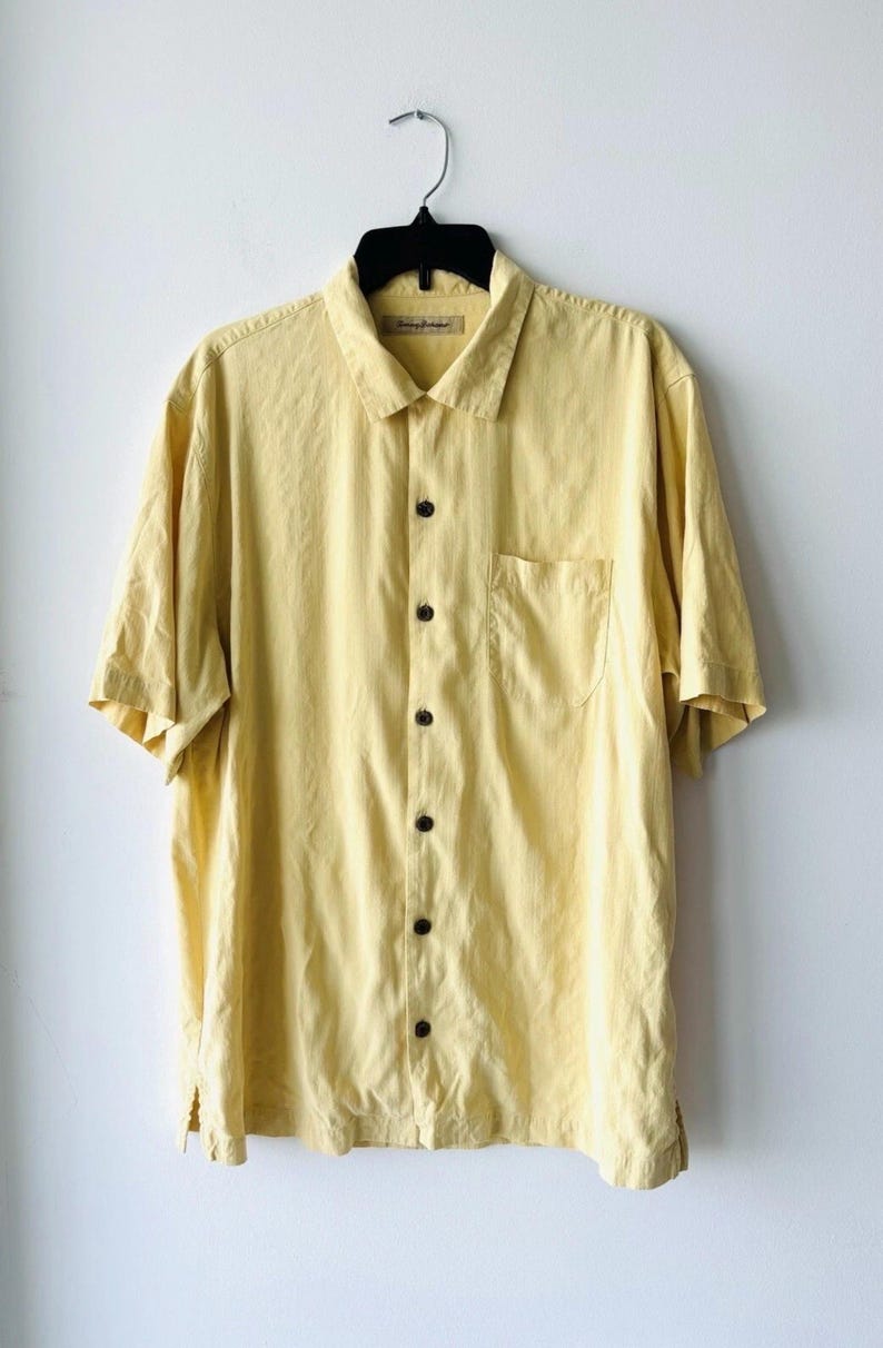 May include: A pale yellow short-sleeved shirt with a button-down front and a collared neckline. It features a single chest pocket and dark brown buttons. The shirt is suspended on a black hanger against a white backdrop.