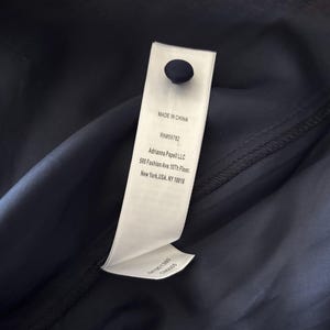May include: A dark navy blue fabric garment featuring a white rectangular clothing label attached with a single dark navy button. The label displays the text: MADE IN CHINA, RN#59782, Adrianna Papell LLC, 500 Fashion Ave. 10th Floor, New York, USA, NY 10018.