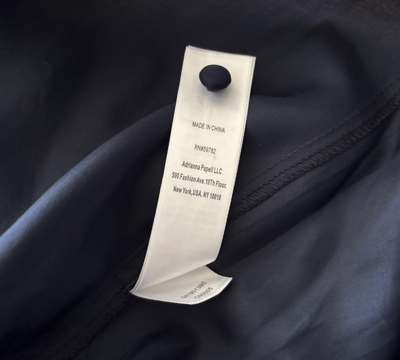 May include: A dark navy blue fabric garment featuring a white rectangular clothing label attached with a single dark navy button. The label displays the text: MADE IN CHINA, RN#59782, Adrianna Papell LLC, 500 Fashion Ave. 10th Floor, New York, USA, NY 10018.