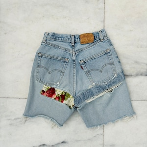 May include: Light blue denim shorts with a strawberry print fabric patch on the back. The shorts have a frayed hem and are distressed.
