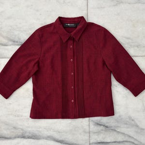 May include: A burgundy-colored blouse with a collared neckline and three-quarter sleeves. The front features vertical pleats and button closures. The fabric has a subtle texture, and the blouse is laid flat on a marble surface.