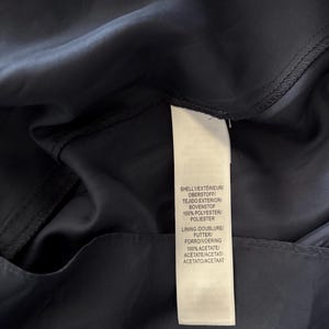 May include: A dark navy blue garment featuring a white fabric care label. The label text reads: SHELL 1/EXT&Eacute;RIEUR/OBERSTOFF/TEJIDO EXTERIOR/BOVENSTOF 100% POLYESTER/POLIESTER and LINING/DOUBLURE/FUTTER/FORRO/VOERING 100% ACETATE/AC&Eacute;TATE/ACETAT/ACETATO/ACETAAT.