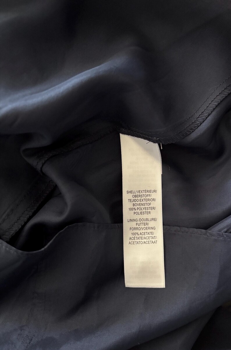 May include: A dark navy blue garment featuring a white fabric care label. The label text reads: SHELL 1/EXT&Eacute;RIEUR/OBERSTOFF/TEJIDO EXTERIOR/BOVENSTOF 100% POLYESTER/POLIESTER and LINING/DOUBLURE/FUTTER/FORRO/VOERING 100% ACETATE/AC&Eacute;TATE/ACETAT/ACETATO/ACETAAT.
