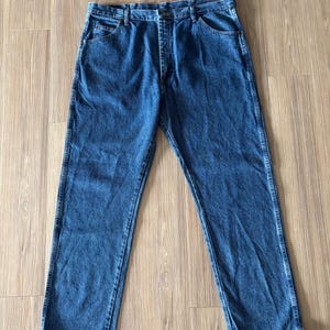 May include: A pair of blue denim jeans with a classic five-pocket design. The jeans have a straight leg cut and a button and zipper closure. The denim is a medium wash, and the jeans are laid flat on a wooden surface.