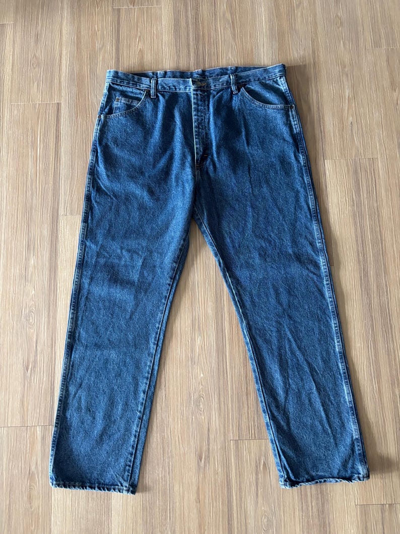 May include: A pair of blue denim jeans with a classic five-pocket design. The jeans have a straight leg cut and a button and zipper closure. The denim is a medium wash, and the jeans are laid flat on a wooden surface.
