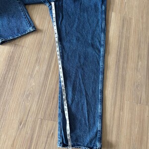 May include: Blue denim jeans with a straight leg design. A measuring tape shows the length of the jeans. The jeans are laid out on a light brown wooden surface.