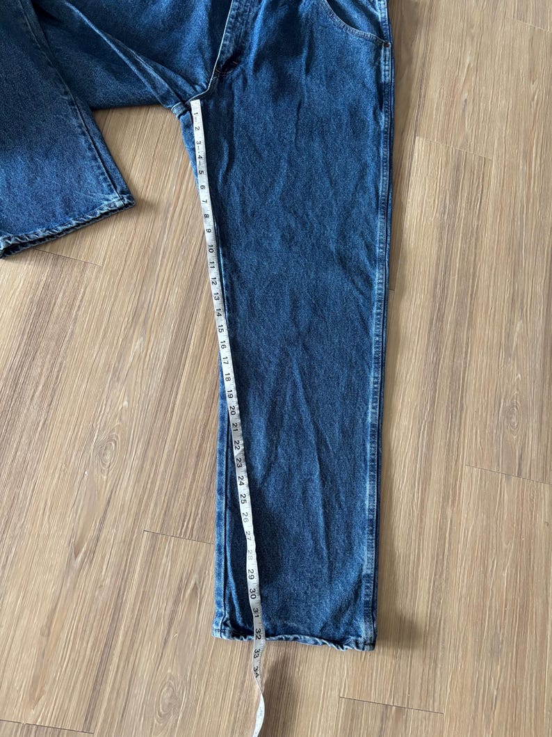 May include: Blue denim jeans with a straight leg design. A measuring tape shows the length of the jeans. The jeans are laid out on a light brown wooden surface.