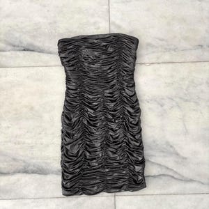 May include: A strapless, ruched mini dress in a dark, metallic gray. The dress features a fitted silhouette with intricate gathering details throughout the fabric, creating a textured appearance. The dress is displayed on a marble surface.