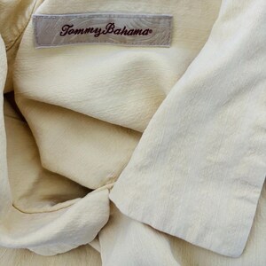 May include: Close-up of a pale yellow shirt with the label "Tommy Bahama" visible. The shirt has a textured fabric and a folded collar. The label is rectangular with the brand name in a cursive font.