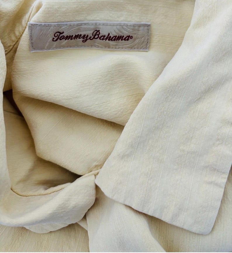 May include: Close-up of a pale yellow shirt with the label "Tommy Bahama" visible. The shirt has a textured fabric and a folded collar. The label is rectangular with the brand name in a cursive font.