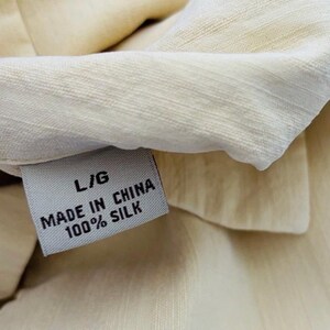 May include: Close-up of a cream-coloured silk garment with a label that reads "L/G MADE IN CHINA 100% SILK." The fabric has a subtle texture and sheen. The label is white with black text.